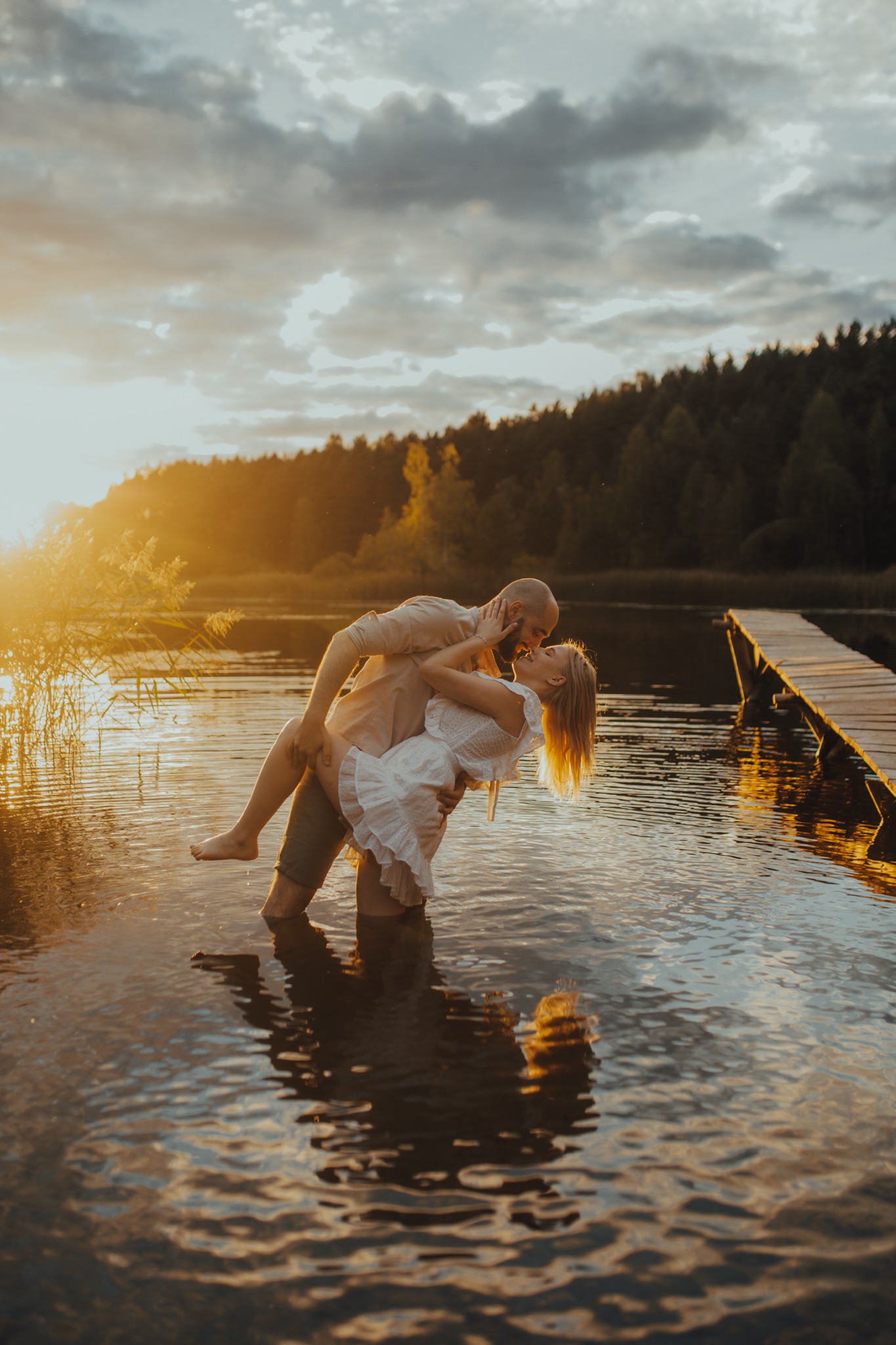 Para w objęciach w wodzie jeziora o zachodzie słońca, drewniany pomost w tle, ciepłe oświetlenie.