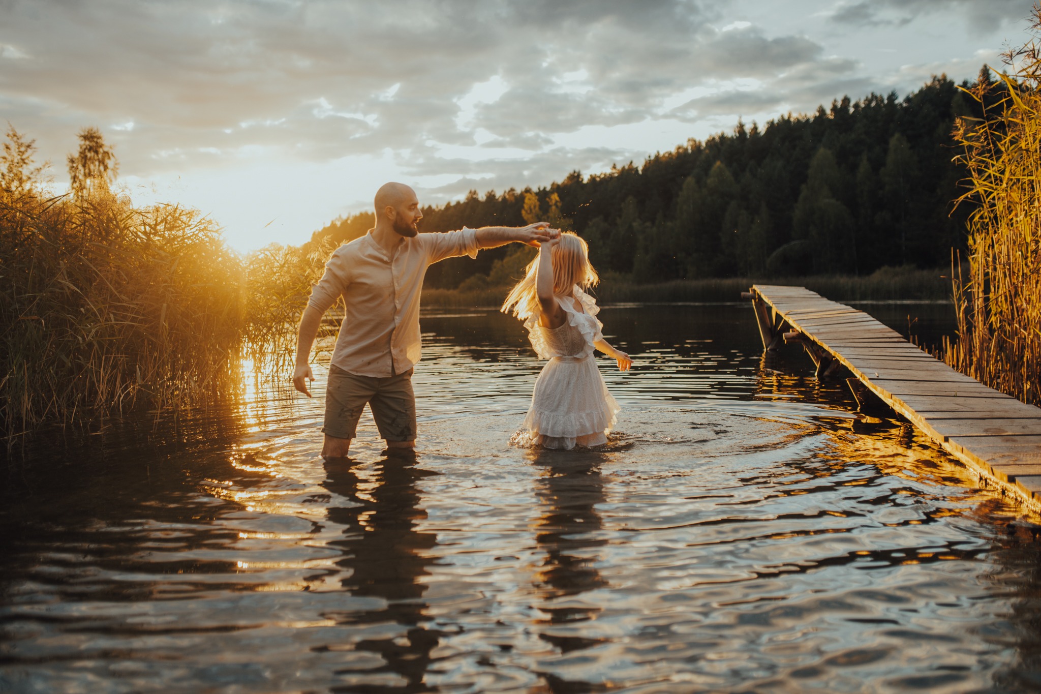 Para młoda tańczy w jeziorze o zachodzie słońca, drewniany pomost w tle, trzciny i las na horyzoncie.