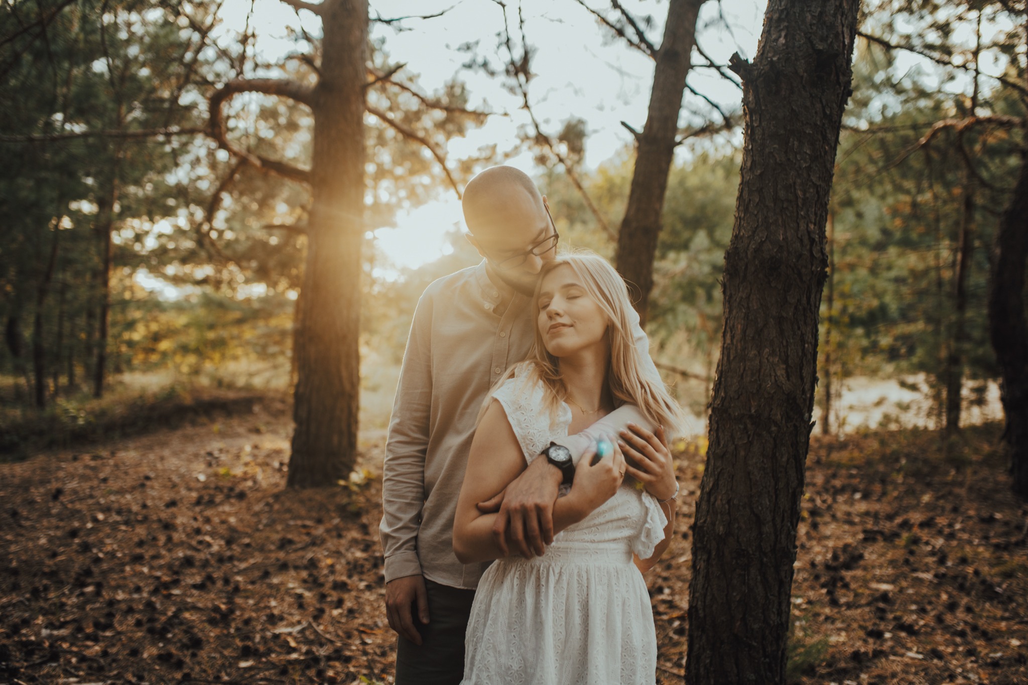 Zakochana para w lesie: mężczyzna w okularach obejmuje blondynkę w białej sukience, ciepłe promienie słońca w tle.