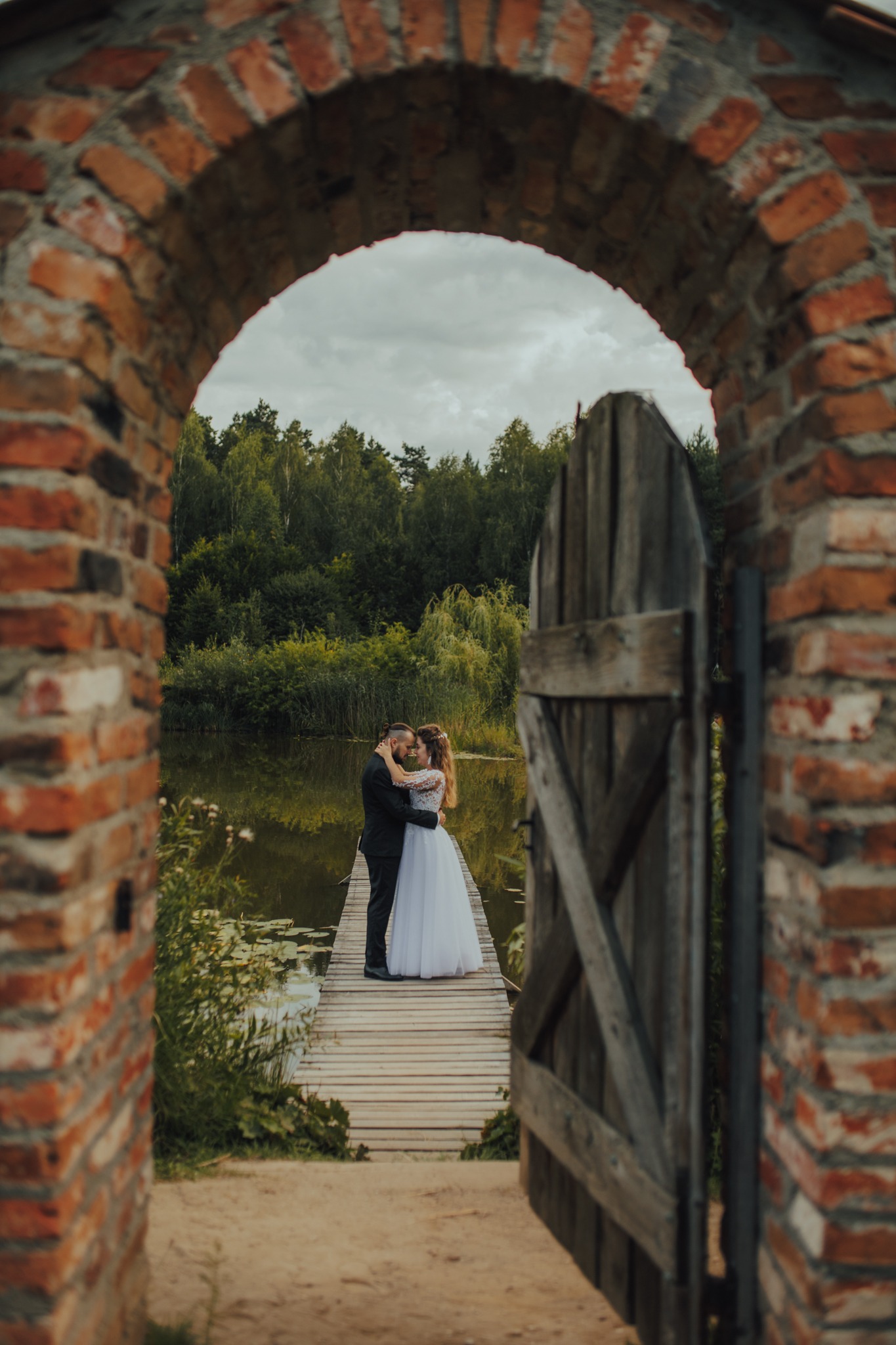 Para młoda w objęciach na drewnianym pomoście nad jeziorem, widziana przez ceglany łuk i drewnianą furtkę.