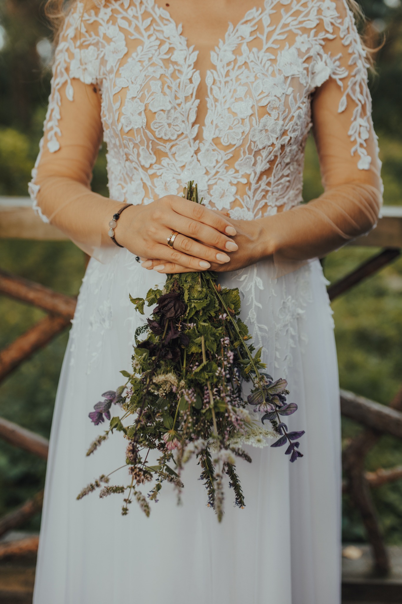 Panna młoda w koronkowej sukni trzyma bukiet polnych kwiatów z fioletowymi akcentami, widoczny złoty pierścionek na palcu serdecznym.