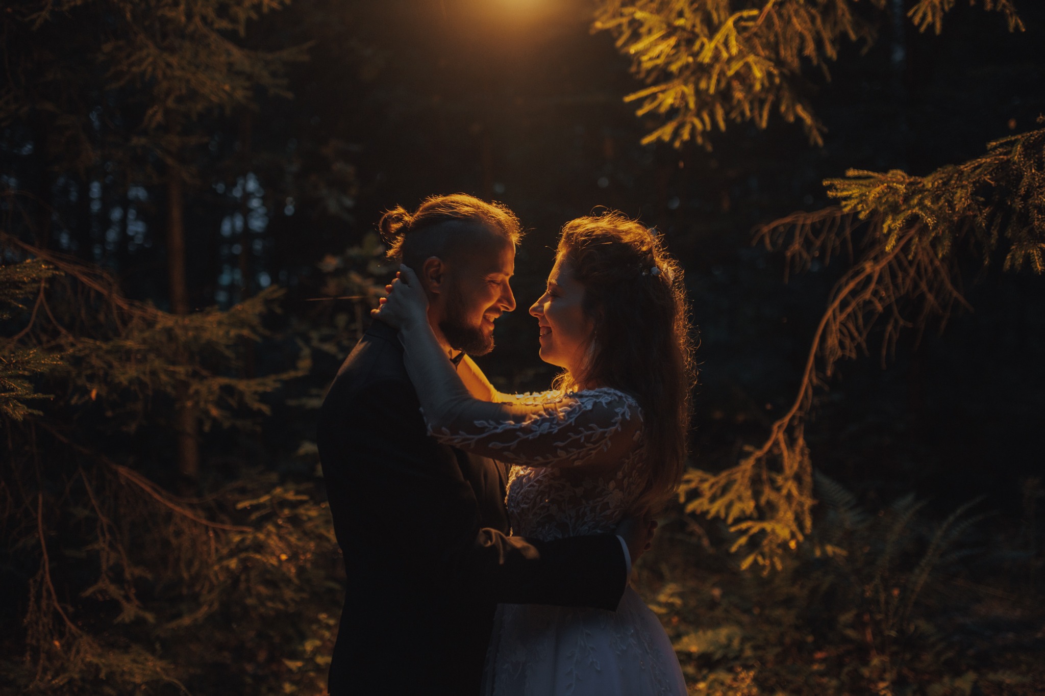Uśmiechnięta para młoda w objęciach, on w czarnym garniturze z muchą, ona w białej sukni z koronkowymi rękawami, oświetleni ciepłym, punktowym światłem na tle ciemnego lasu.