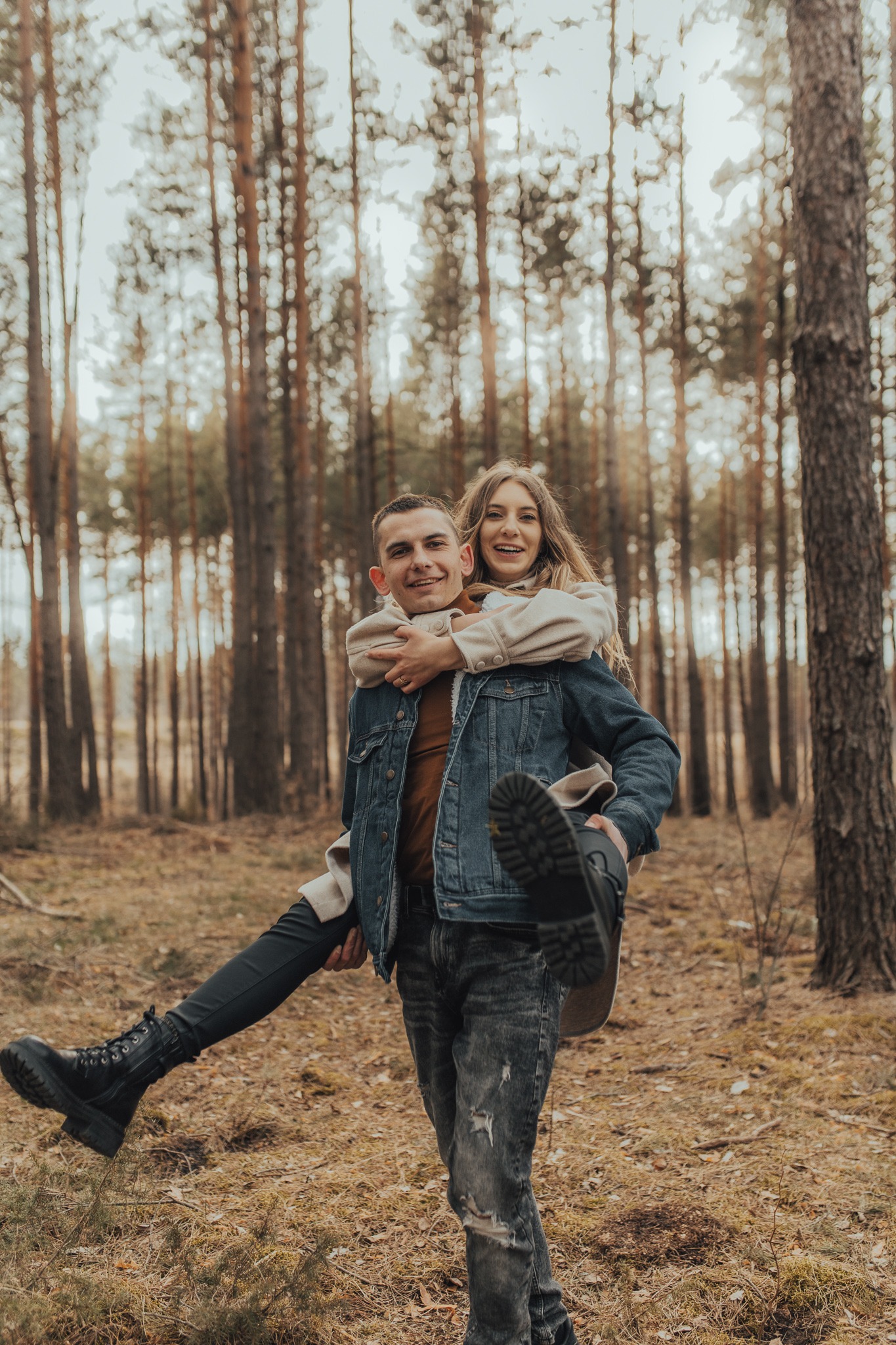 Uśmiechnięta para w lesie: mężczyzna trzyma na barana kobietę w jasnej kurtce, widoczne wysokie sznurowane buty i jeansowe kurtki.