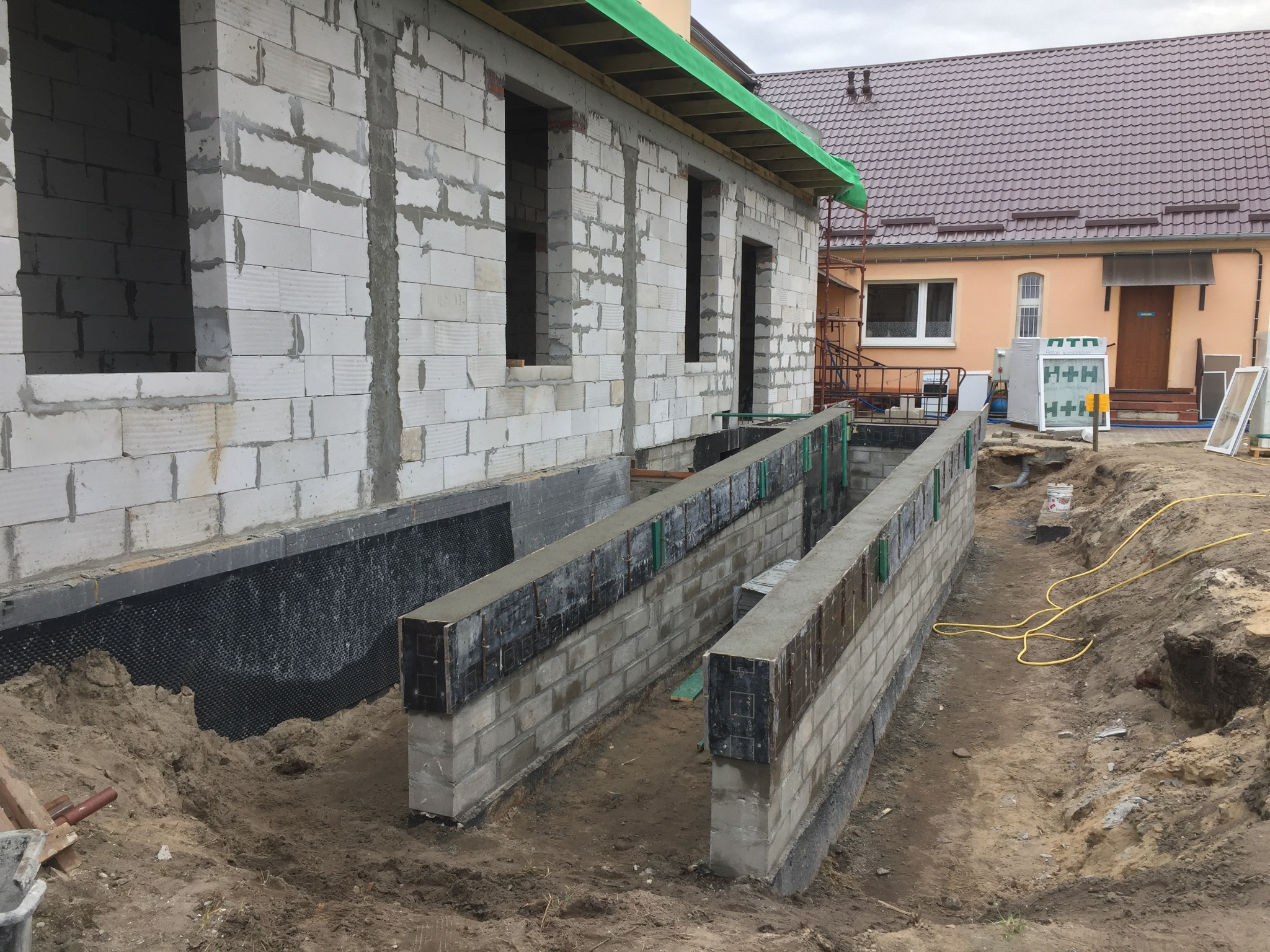 Budowa domu z bloczków: wznoszone ściany fundamentowe i ściany parteru, widoczne okna bez ram, zabezpieczenie przeciwwilgociowe fundamentów, teren budowy.