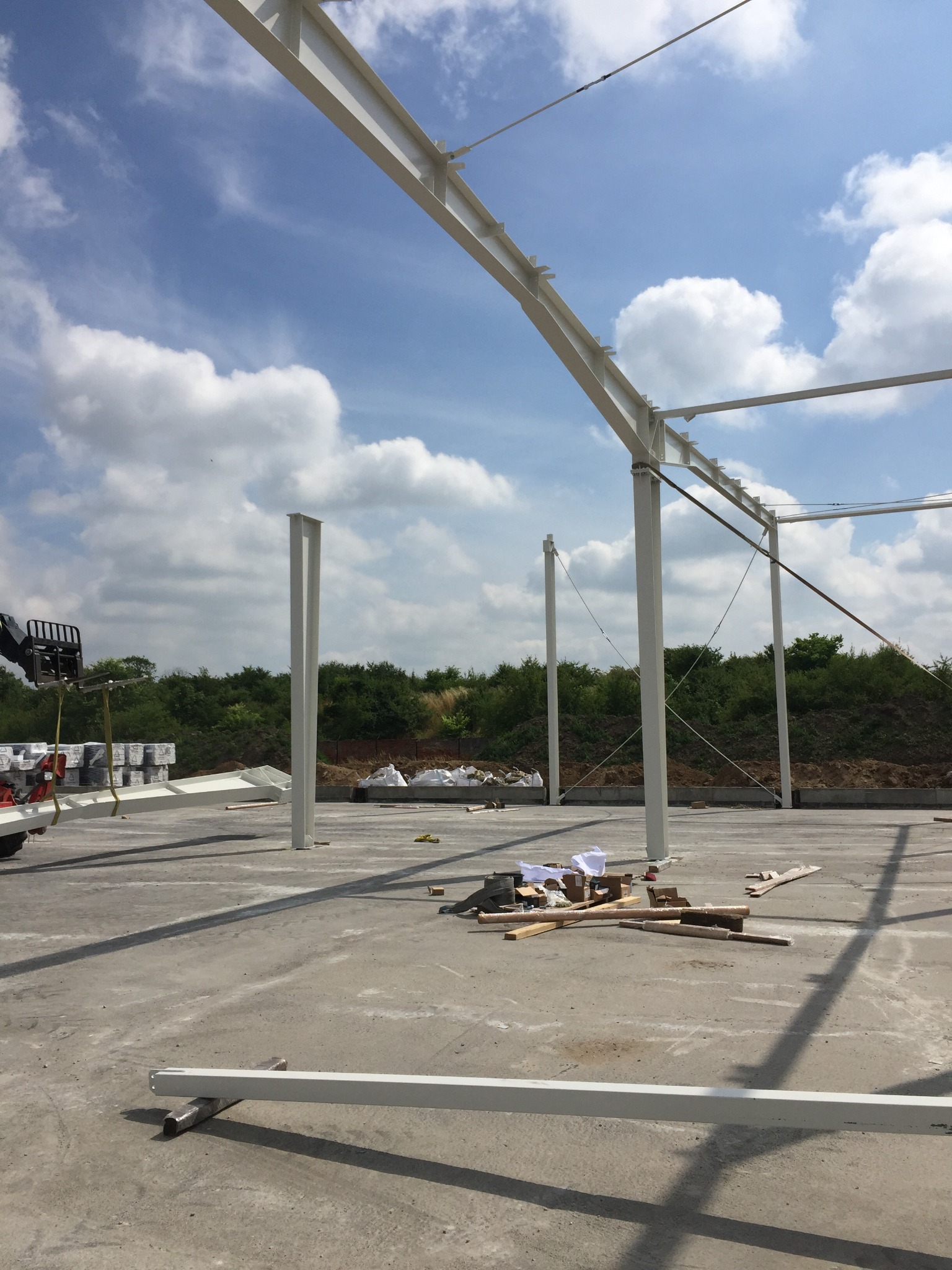 Montaż białej konstrukcji stalowej hali na betonowej płycie fundamentowej, widoczny wózek widłowy z elementami konstrukcyjnymi, błękitne niebo z chmurami w tle.