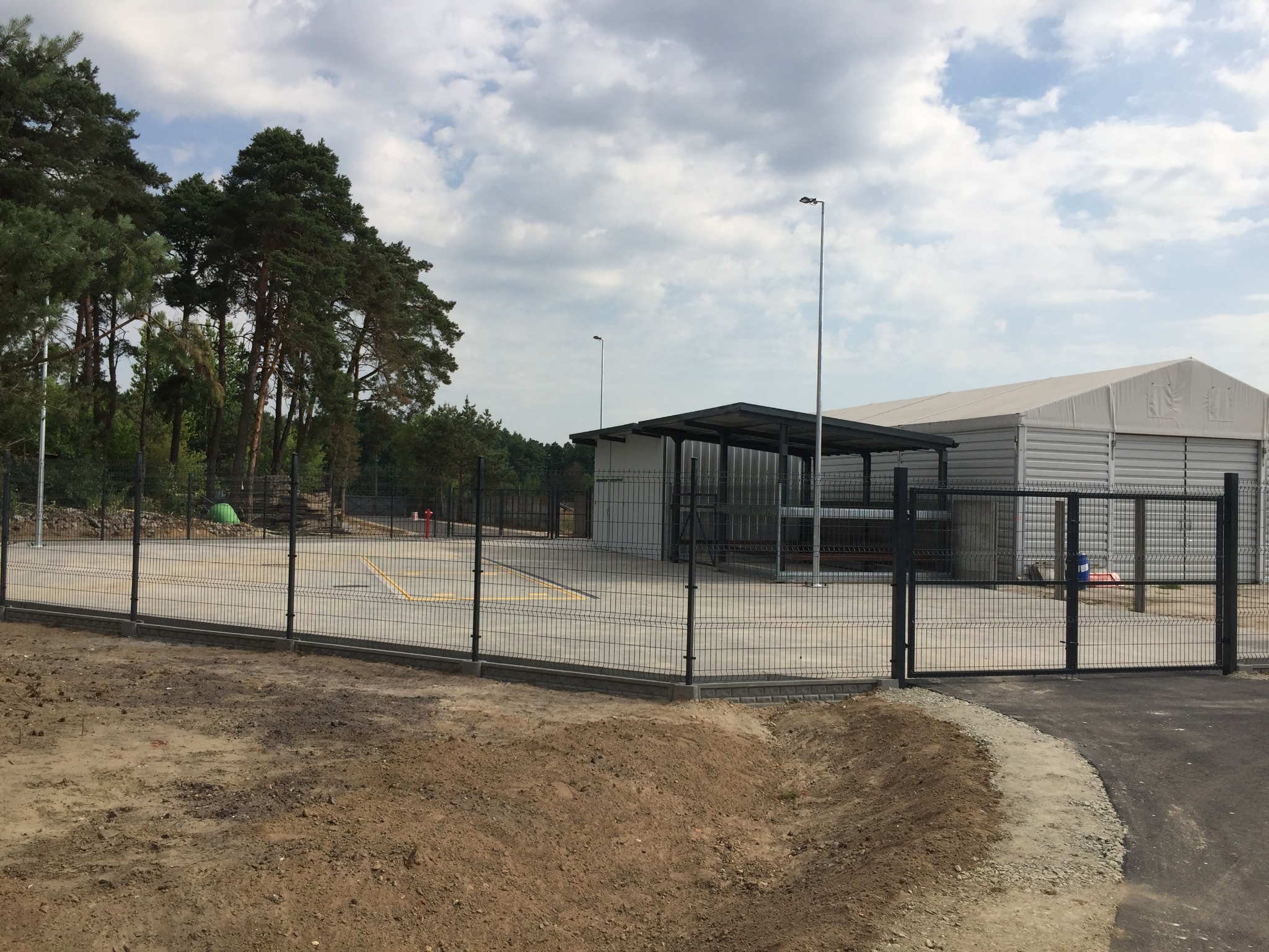 Teren ogrodzony siatką panelową, z widoczną w tle konstrukcją wiaty i hali namiotowej, otoczony zielenią drzew.