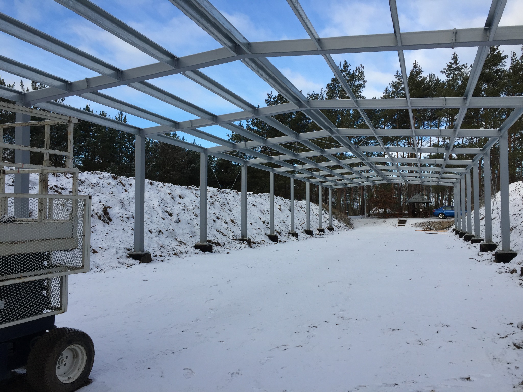 Konstrukcja szkieletowa budynku w trakcie montażu, widoczne stalowe słupy i belki, na ziemi śnieg, w tle las.