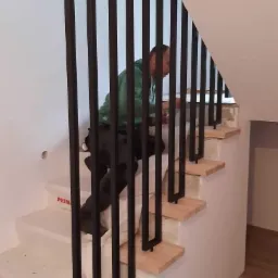 Montaż czarnych, metalowych balustrad pionowych na drewnianych stopniach schodów, widoczny pracownik w trakcie instalacji.