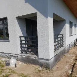 Nowoczesny dom z szarą elewacją i ciemnymi, metalowymi balustradami balkonowymi o poziomym układzie szczebli. Widoczny wykop wzdłuż fundamentów i żółty wąż ogrodowy.