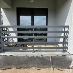 Szara, pozioma balustrada metalowa na balkonie, widoczne narzędzia montażowe i okno z odbiciem nieba.