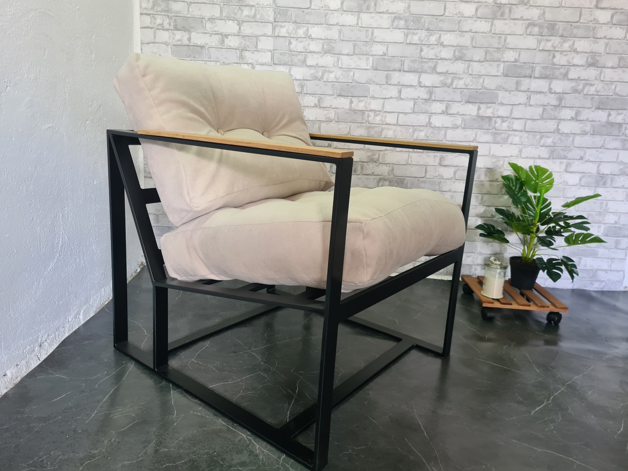 Nowoczesny fotel z czarną metalową ramą i beżowymi poduszkami na tle ściany z białej cegły i rośliny doniczkowej.