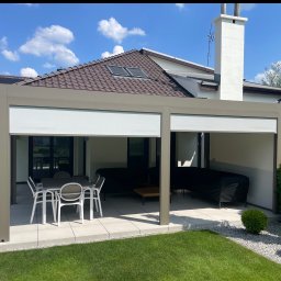 Pat-Mar Gliwice Sp. z o.o. - Nowoczesna pergola tarasowa z beżową konstrukcją i zwijanymi roletami, ustawiona na jasnym tarasie z meblami ogrodowymi, na tle domu z brązowym dachem.
