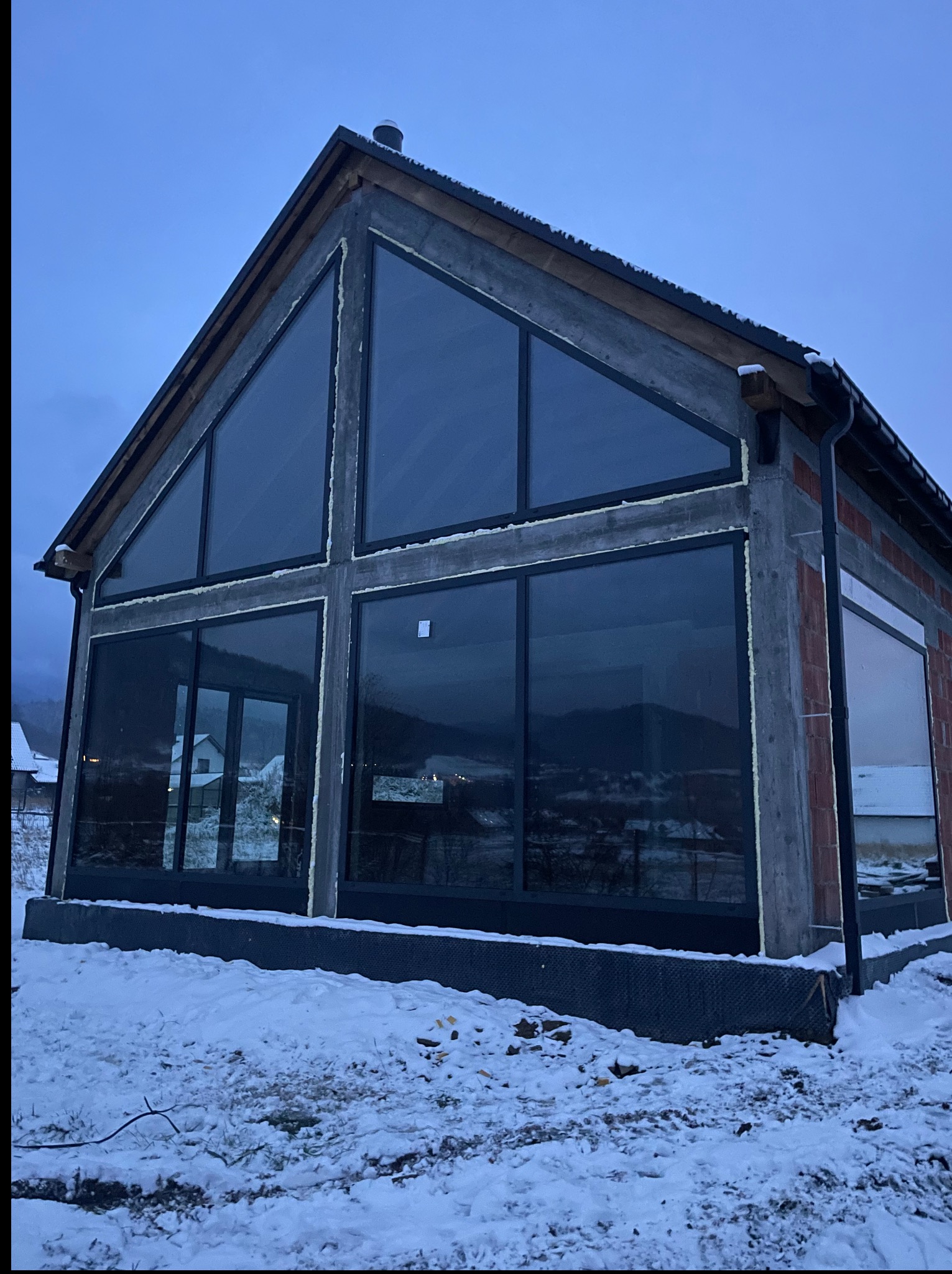 Dom w stanie surowym z dużymi, panoramicznymi oknami z czarnymi ramami. Widoczne ocieplenie pianką montażową. Zimowy krajobraz w tle.