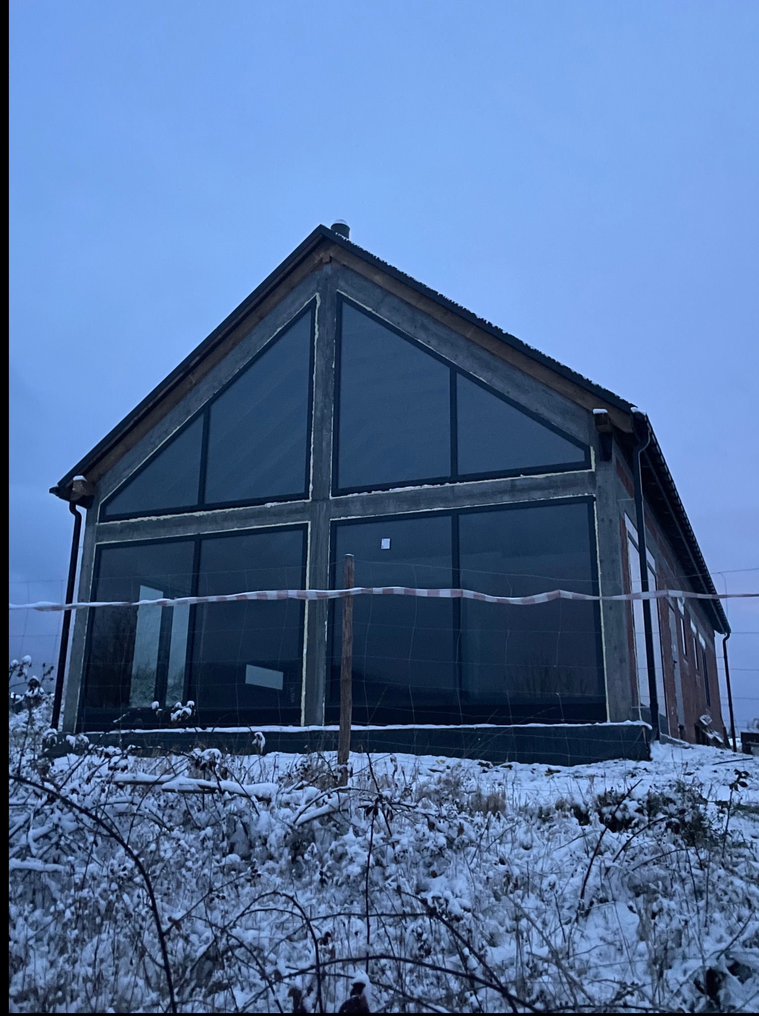 Dom w budowie z dużymi oknami w kształcie trójkąta i prostokątów, otoczony śniegiem. Widoczne zabezpieczenia budowlane.