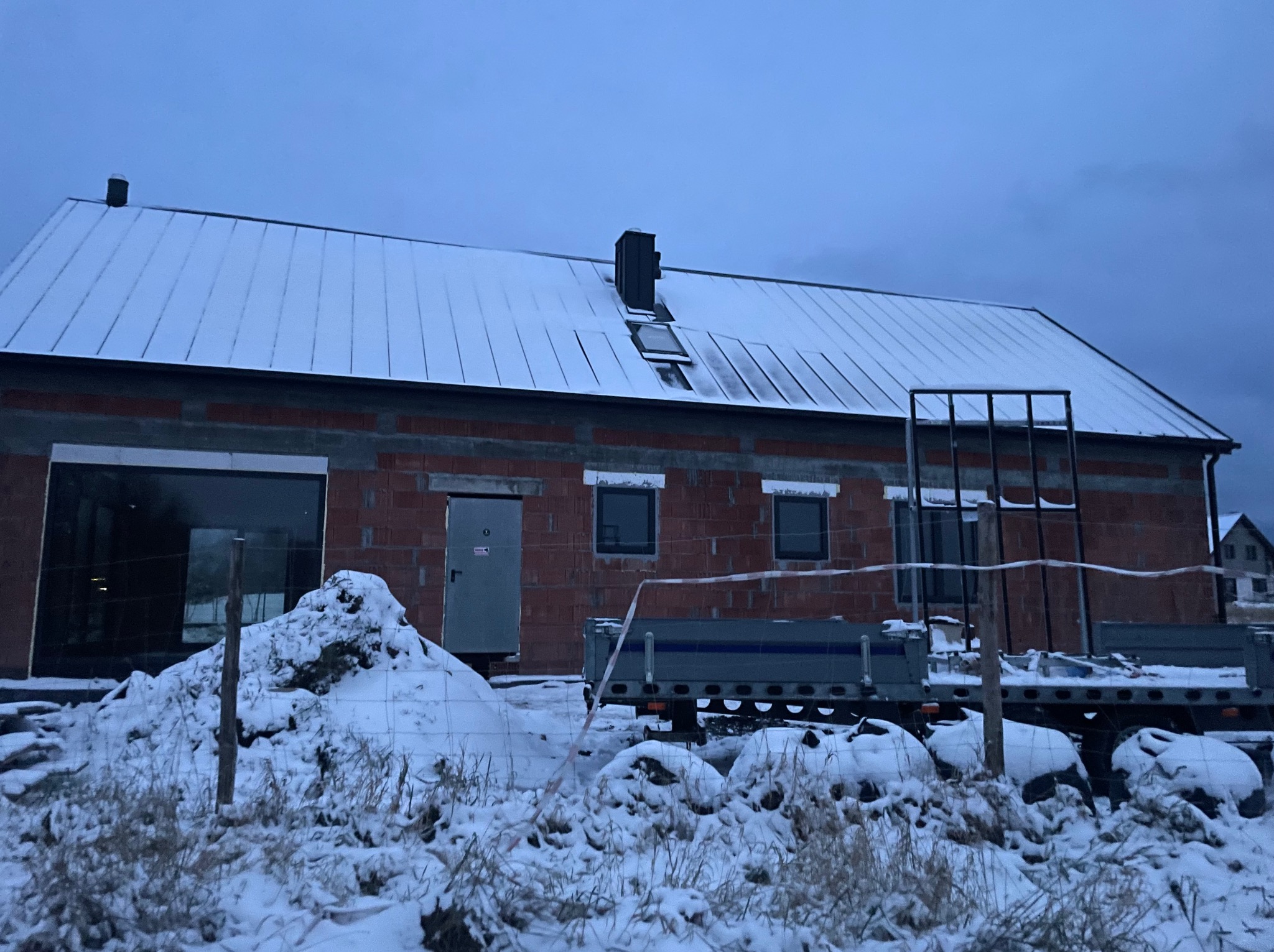 Surowy budynek w zimowej scenerii: czerwona cegła, okna, metalowe drzwi, dach pokryty śniegiem, przyczepa i zaspy śnieżne. Ujęcie zza ogrodzenia.