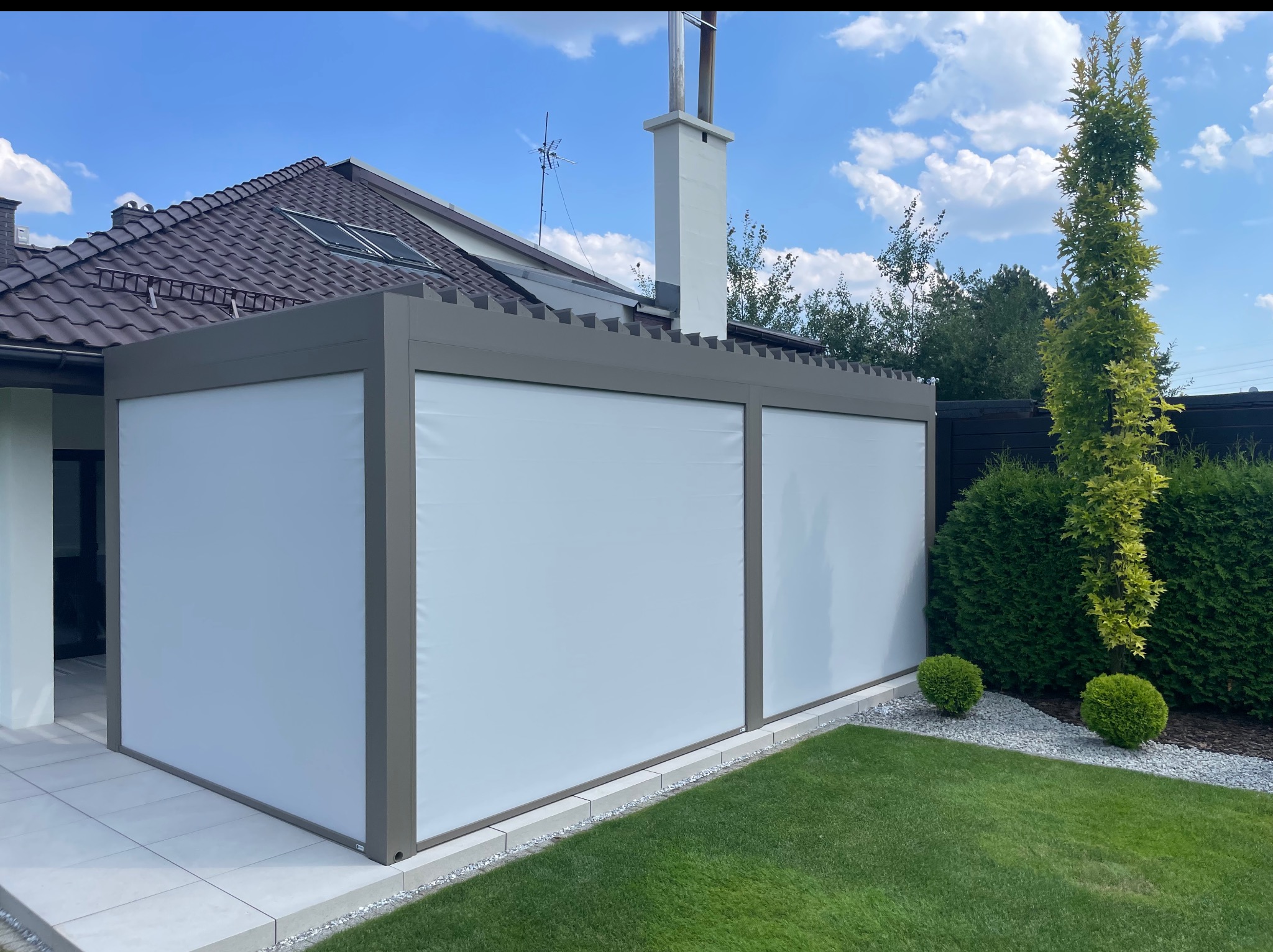 Nowoczesna pergola z białymi, zwijanymi osłonami przeciwsłonecznymi, na tle domu z brązowym dachem i zielonego trawnika w słoneczny dzień.