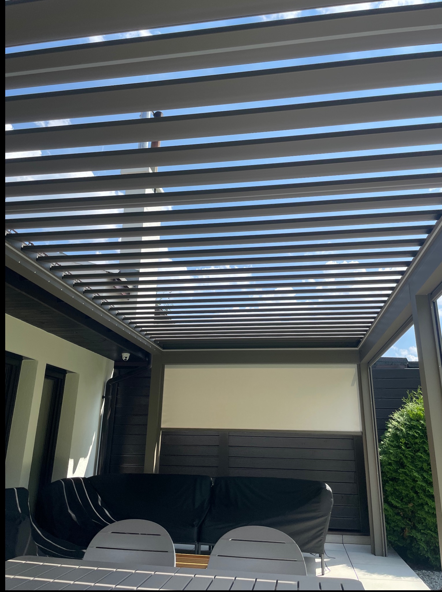 Nowoczesna pergola tarasowa z regulowanymi lamelami, widok z dołu na błękitne niebo. Minimalistyczny design, szare profile, oświetlenie punktowe. Część mebli ogrodowych.