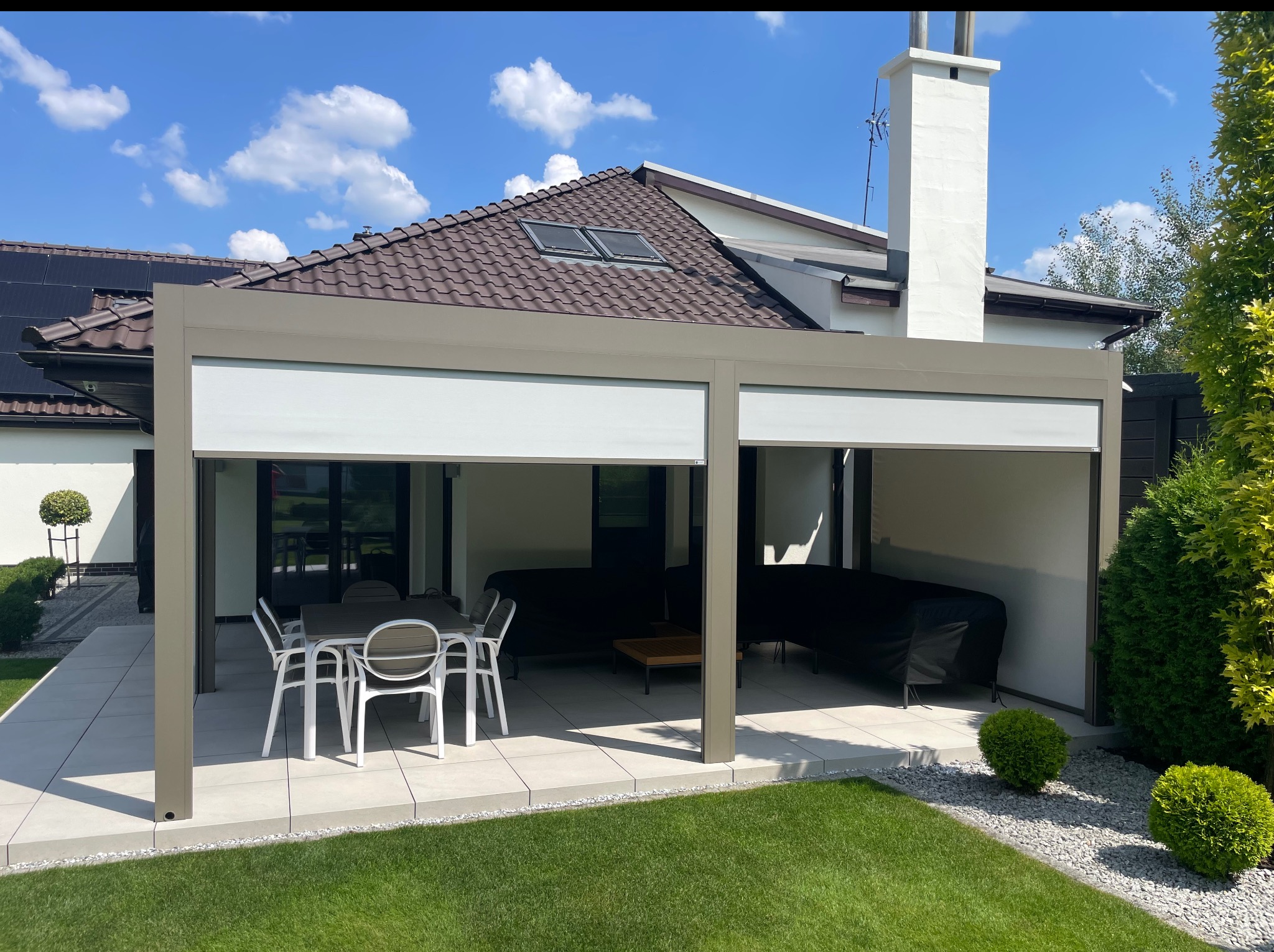 Nowoczesna pergola tarasowa z beżową konstrukcją i zwijanymi roletami, ustawiona na jasnym tarasie z meblami ogrodowymi, na tle domu z brązowym dachem.