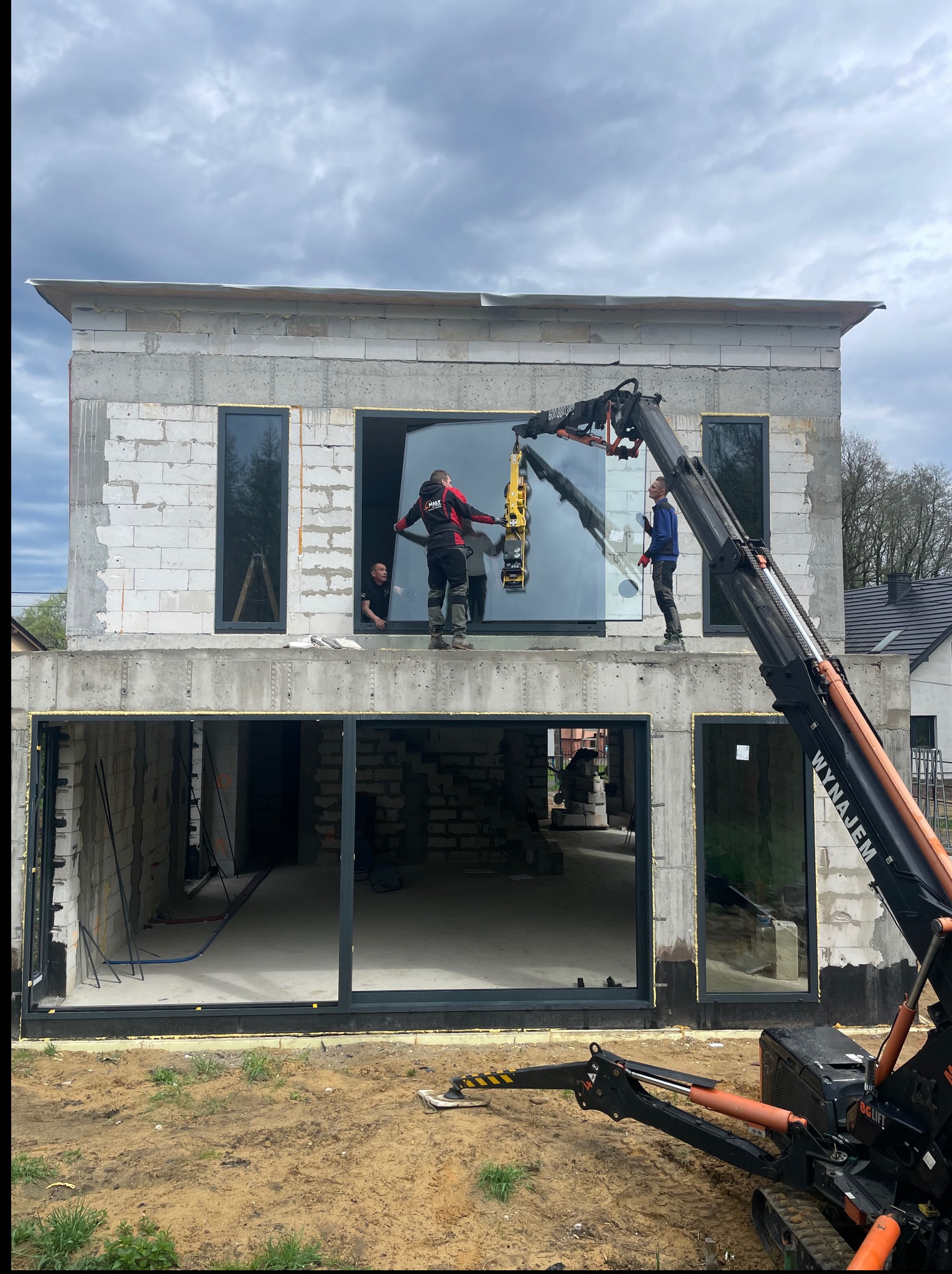 Montaż dużego okna przez ekipę budowlaną za pomocą dźwigu na placu budowy domu jednorodzinnego w stanie surowym otwartym. Widoczne czarne ramy okienne.