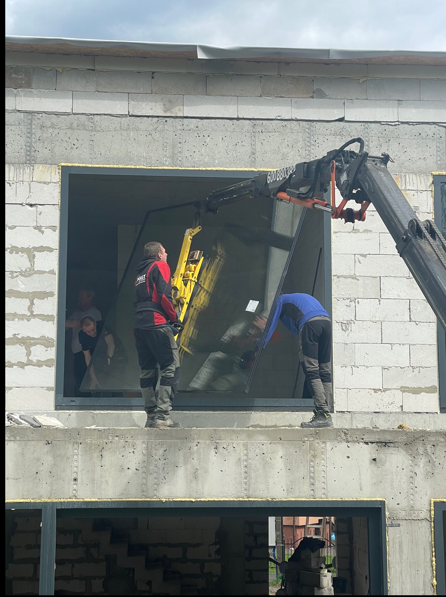 Montaż okna na budowie z użyciem dźwigu. Dwóch monterów przy oknie, w tle widoczni inni ludzie. Surowy stan budynku z betonu komórkowego.