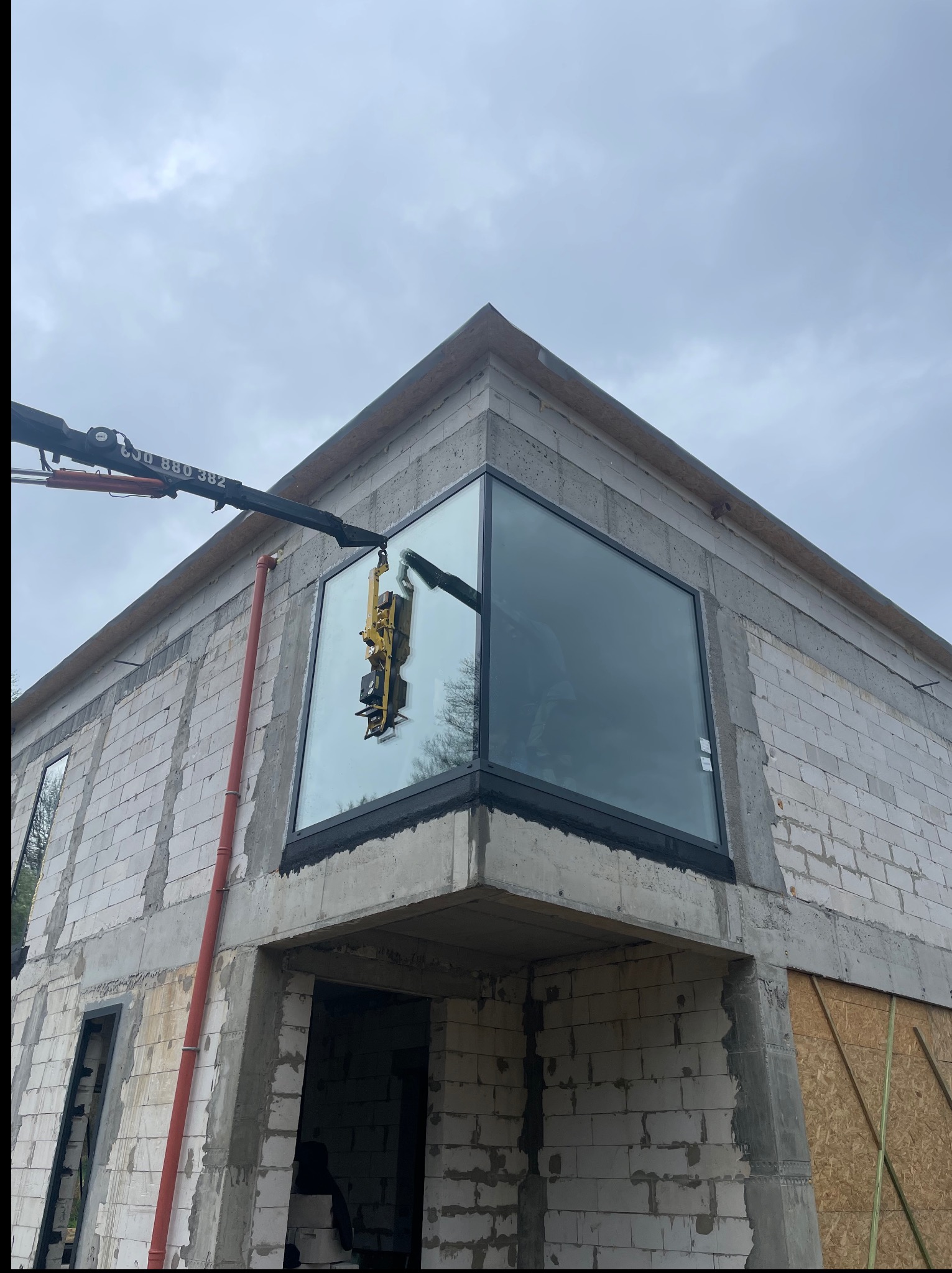 Montaż dużego okna na surowej ścianie budynku za pomocą przyssawki na wysięgniku dźwigu. Widoczne bloczki betonowe i fragment instalacji.