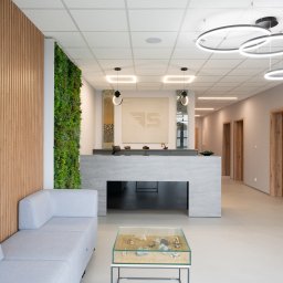 Boska Design - Nowoczesna recepcja w Opolu z betonową ladą, zieloną ścianą wertykalną, drewnianymi lamelami i szklanym stolikiem z miniaturowymi maszynami budowlanymi. Jasne wnętrze z designerskim oświetleniem.