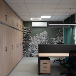 Boska Design - Nowoczesne biuro z długą ścianą drewnianych szafek z kluczami, ciemnozieloną ścianą z tapetą w czarno-białe drzewa i minimalistycznym biurkiem z czarną przegrodą.