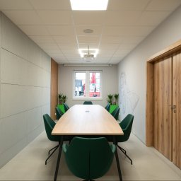 Boska Design - Nowoczesna sala konferencyjna z długim drewnianym stołem i ośmioma zielonymi krzesłami. Ściany z paneli betonowych, drewnianych lameli i szkicem architektonicznym. Podwieszany sufit z projektorem.