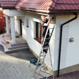 Instalator na drabinie montuje kamerę monitoringu na elewacji domu z czerwonym dachem i dekoracjami Halloween.