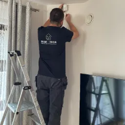 Instalator w czarnym t-shircie z logo firmy WOB-TECH montuje czujnik na ścianie w rogu pokoju, stojąc na aluminiowej drabinie Krause, widoczny fragment telewizora i zasłony w tle.