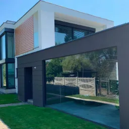 Nowoczesny dom z dużymi oknami i roletami zewnętrznymi, widok z zewnątrz w słoneczny dzień, z elementami drewnianej elewacji i szklaną konstrukcją garażu.