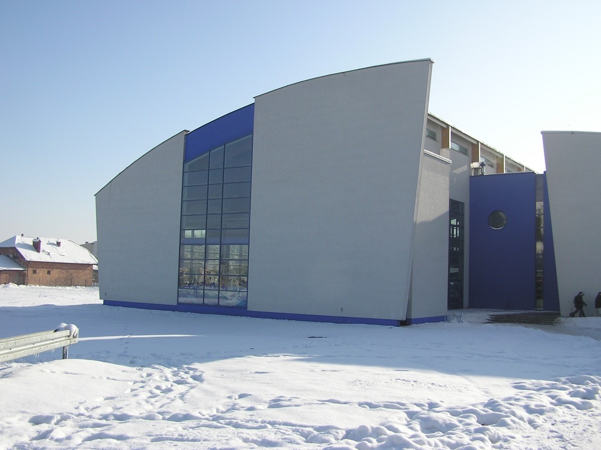 Nowoczesny budynek z dużymi oknami i niebieskimi akcentami architektonicznymi, otoczony śniegiem w zimowy dzień.