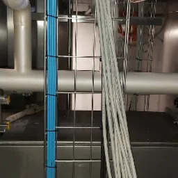 Uporządkowane kable elektryczne w korycie siatkowym, przewody niebieskie i szare spięte opaskami, widoczne rury i elementy instalacji w tle.