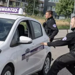 Dwóch ochroniarzy w czarnych mundurach stoi obok białego samochodu z logo firmy ochroniarskiej na dachu i boku pojazdu.