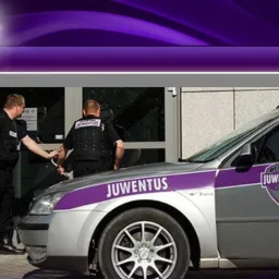 Dwóch ochroniarzy w czarnych uniformach przy srebrnym samochodzie z logo firmy ochroniarskiej 'Ochrona Juwentus Grupa' na tle szklanego wejścia do budynku.