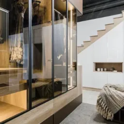 Nowoczesna garderoba z przeszklonymi frontami, oświetleniem LED i drewnianymi elementami, wkomponowana pod schody z białymi szafkami.