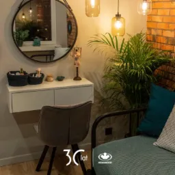 Biała, wisząca toaletka z szufladami, okrągłe lustro w czarnej ramie, szara welurowa krzesło, wiszące lampy w stylu retro, ceglana ściana w tle, roślina doniczkowa, narożnik z poduszkami, logo...
