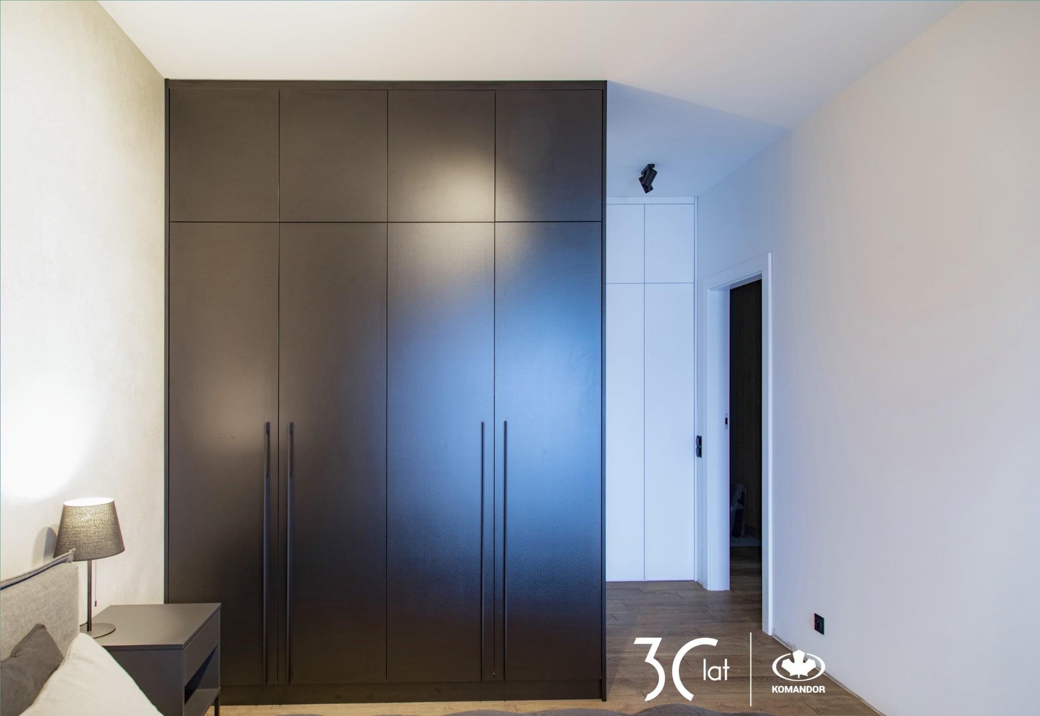 Duża, grafitowa szafa z długimi, pionowymi uchwytami, w sypialni z lampką nocną na stoliku i widocznym fragmentem łóżka. W tle białe drzwi i fragment podłogi z paneli.