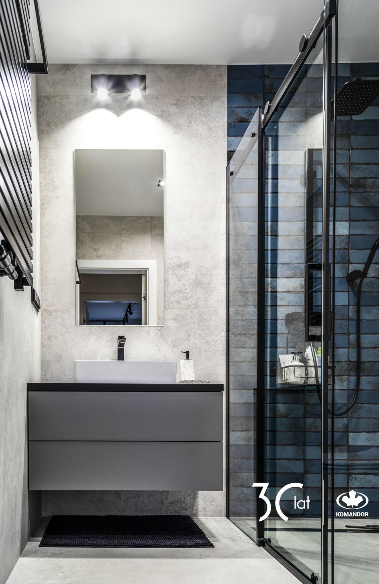 Nowoczesna łazienka z szarą szafką pod umywalką, prostokątnym lustrem z oświetleniem, grzejnikiem drabinkowym i kabiną prysznicową z czarnymi profilami i niebieską mozaiką na ścianie, widoczne logo...