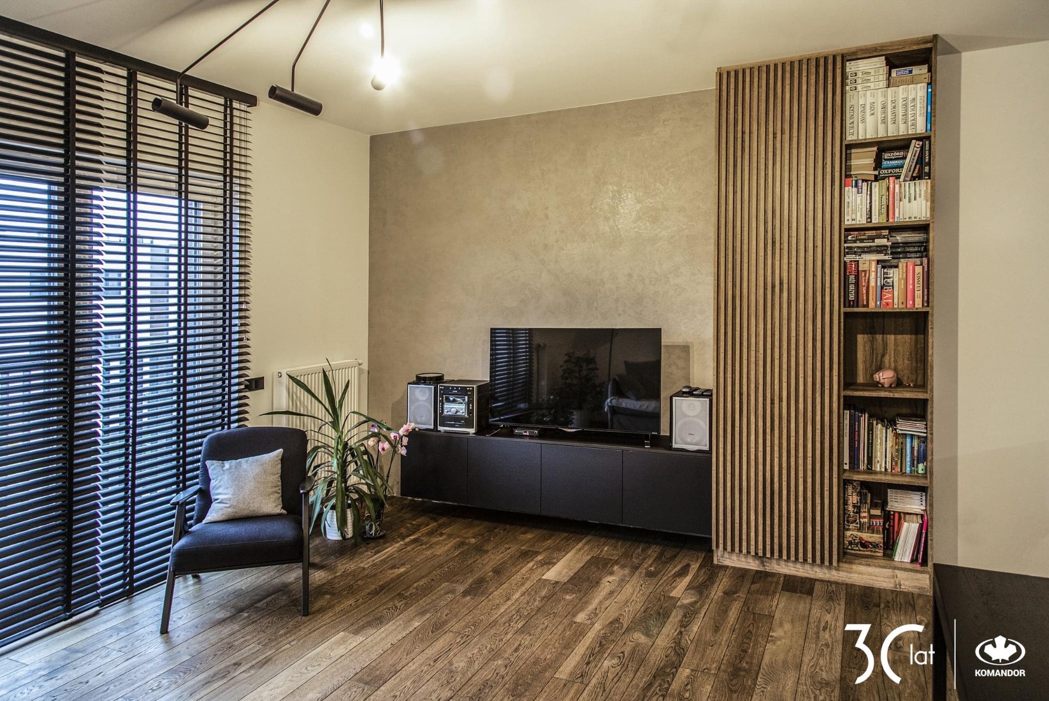 Nowoczesny salon z drewnianą podłogą, czarnymi żaluzjami, szarym fotelem z poduszką, szafką RTV z czarnymi frontami, głośnikami, telewizorem i regałem z drewnianymi listwami oraz książkami.