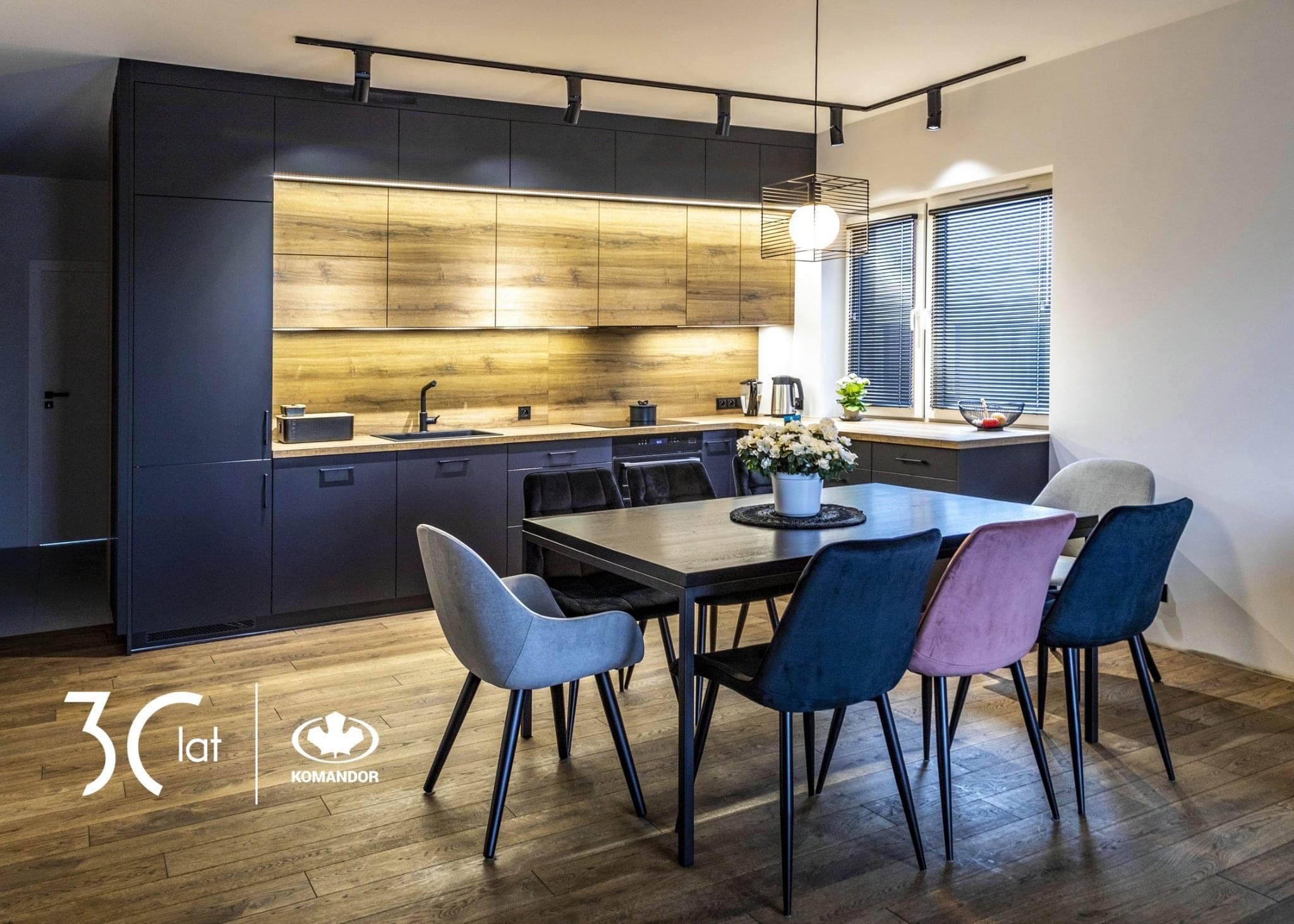 Nowoczesna kuchnia z ciemnymi, matowymi szafkami i drewnianymi panelami, oświetlona listwą LED, z czarnym stołem i kolorowymi krzesłami na drewnianej podłodze; widoczne logo firmy Komandor w rogu