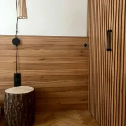 Drewniane panele ścienne w ciepłym odcieniu, z pionowymi listwami na ścianie po prawej stronie i blatem z litego drewna. Obok stoi stołek z pnia drzewa, a nad nim wisi lampa z abażurem z cienkich...