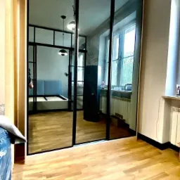 Przesuwne drzwi z czarnymi ramami szprosów, dzielące przestrzeń pokoju z widokiem na sypialnię z łóżkiem i lampą wiszącą, parkiet na podłodze.