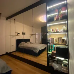 Szafa z lustrzanymi drzwiami przesuwnymi i wbudowanym regałem z oświetleniem LED, odbijająca wnętrze sypialni z łóżkiem i drewnianą podłogą.