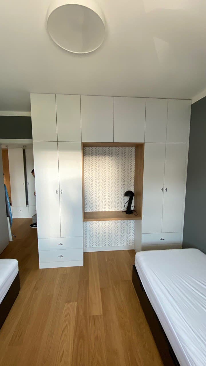 Biała zabudowa szaf wnękowych z drewnianą wnęką i półką, wzorzysta tapeta wewnątrz wnęki, lampka na półce, pokój z dwoma łóżkami.