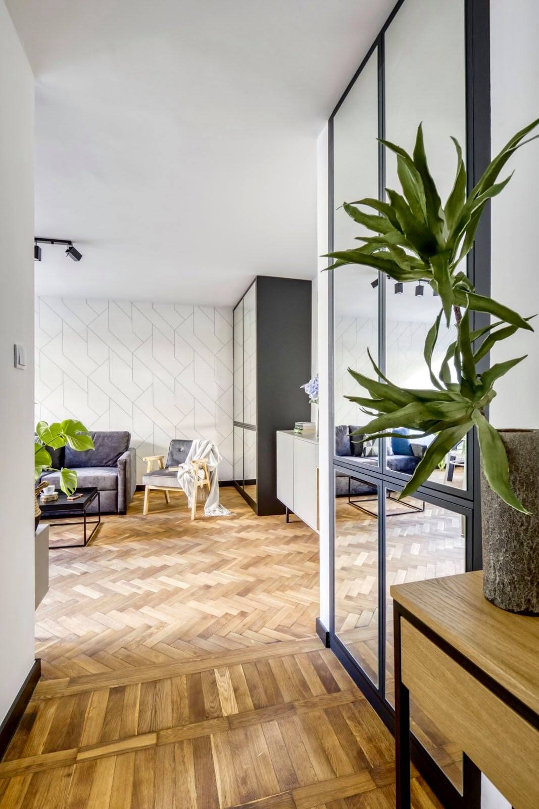 Salon z parkietem ułożonym w jodełkę, szarą sofą, fotel z narzuconym pledem, szafa z lustrzanymi drzwiami i przeszklona ściana działowa z czarnymi szprosami.