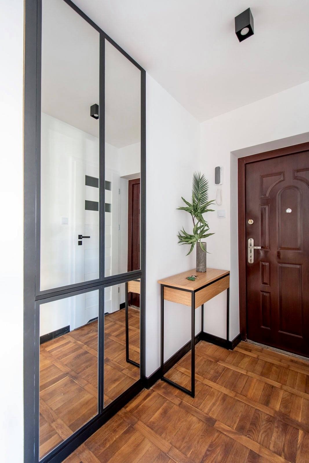 Przeszklona ścianka działowa z czarnymi profilami w przedpokoju w Łomiankach, odbijająca parkiet i drzwi, obok minimalistyczna konsola z drewnianym blatem i rośliną.