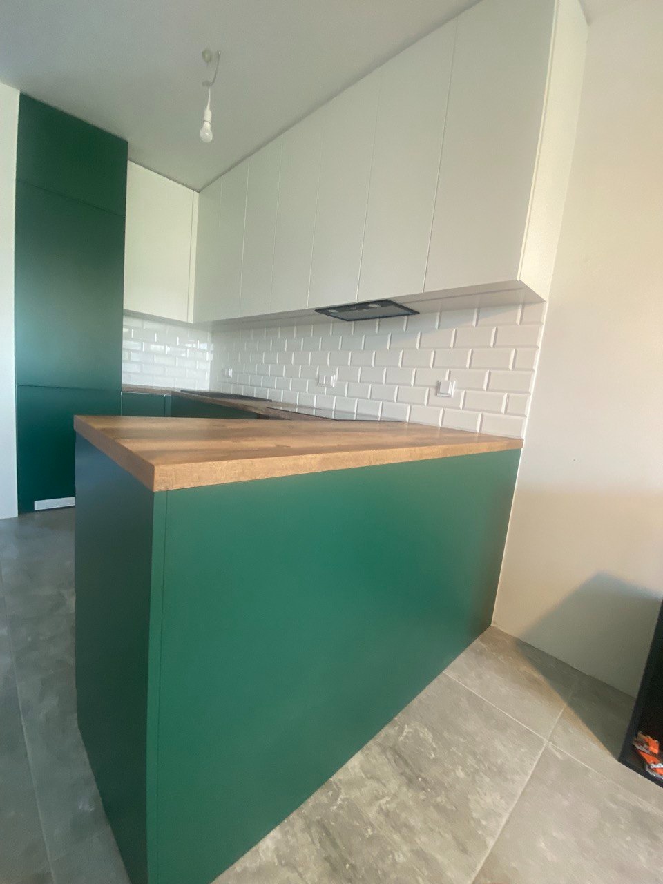 Nowoczesna kuchnia z zielonymi szafkami dolnymi, drewnianym blatem i białymi szafkami górnymi, widok na wyspę kuchenną i fragment ściany z białymi płytkami.