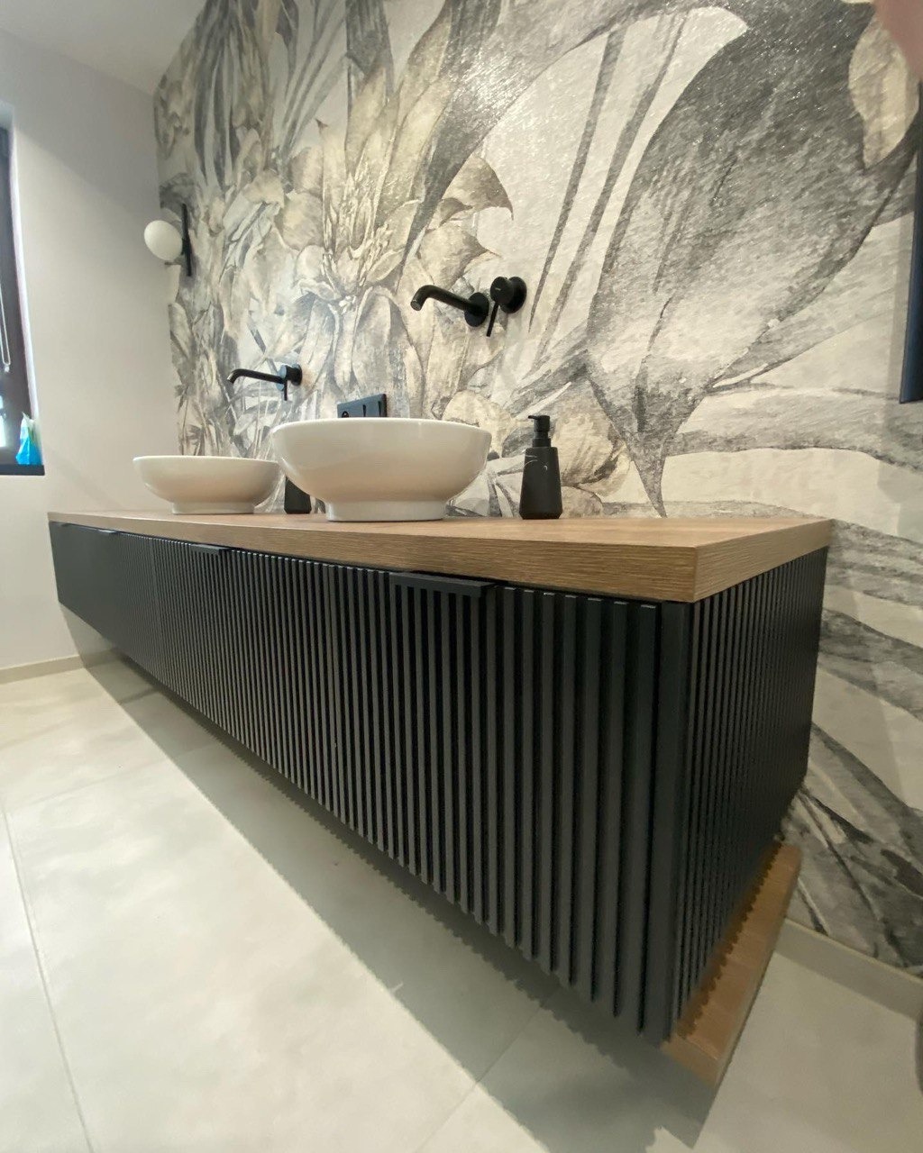 Nowoczesna łazienka z podwieszaną szafką umywalkową z czarnymi, pionowymi lamelami i blatem z jasnego drewna, dwie białe umywalki nablatowe i czarna armatura na tle ściany z tapetą w motywy roślinne.