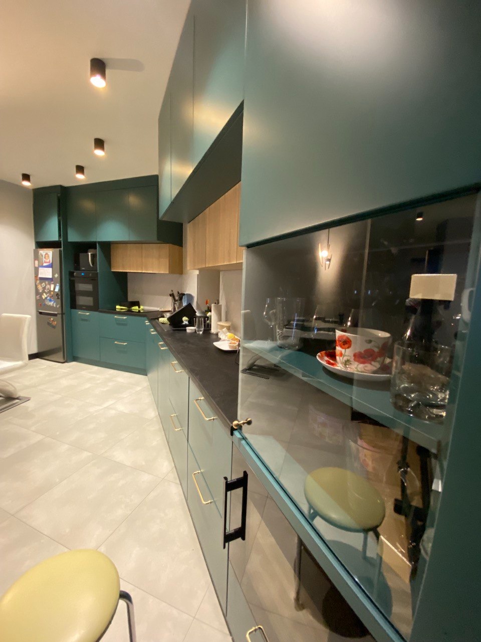 Nowoczesna kuchnia w odcieniach zieleni z czarnym blatem, złotymi uchwytami i przeszklonymi szafkami, oświetlona punktowymi lampami sufitowymi.